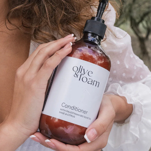 Person holding a bottle of 'Olive & Foam' conditioner with a blurred outdoor background