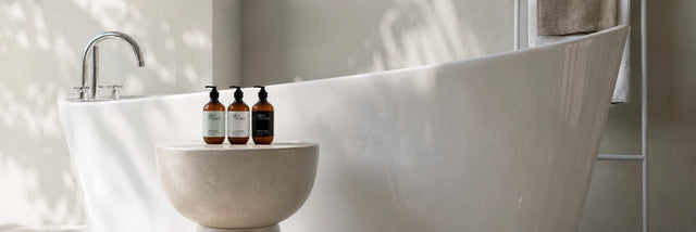 Olive & Foam Shampoo, Conditioner and Body Wash set in amber glass bottles, styled on a stone bowl beside a freestanding bathtub — Palestinian bio olive oil hair and body care